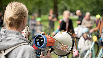 Das Bild zeigt Anika Meenken vom VCD von hinten, wie sie ein Megafon in der Hand hält und zu den Teilnehmenden einer Kidical Mass spricht. Darunter Kinder mit ihren Eltern auf Fahrrädern. Im Hintergrund ist eine Parkanlage mit Bäumen auszumachen.