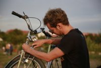 Das Bild zeigt einen jungen Mann mit Sonnenbrille, der im Abendlicht an seinem alten Fahrrad schraubt.