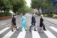vier Menschen gehen mit zusammengeklappten Fahrrädern über einen Zebra-Streifen, ähnlich wie das berühmte Beatles-Cover
