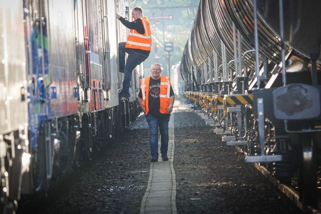 Ein Mann in einer orangefarbenen Warnweste läuft durch einen Korridor aus zwei Güterzügen. Ein zweiter Mann in einer orangefarbenen Warnweste steigt aus einem der Güterzuge aus. 