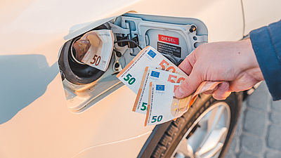 Foto: Hand steckt Geldscheine in den Tankstutzen eines Autos als Symbol für hohe Kraftstoffkosten.
