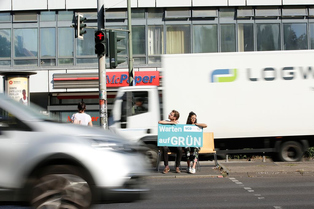 Zwei Personen sitzen auf einer Mittelinsel zwischen zwei mehrspurigen Straße und halten ein Schild in den Händen auf dem steht "Warten auf GRÜN"