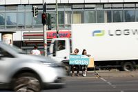 Zwei Personen sitzen auf einer Mittelinsel zwischen zwei mehrspurigen Straße und halten ein Schild in den Händen auf dem steht "Warten auf GRÜN"