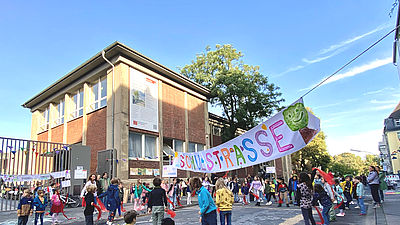 Foto: Schulstraße mit Kindern und Erwachsenen vor einem Schulgebäude, eine bunte Absperrung hält den Autoverkehr fern.