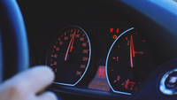 Das Bild zeigt eine Hand am Lenkrad sowie den dahinterliegenden Autotacho, der eine Geschwindigkeit von 130 km/h anzeigt. 