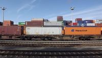 Container auf einem Güterwaggon, im Hintergrund sind weitere Container zu sehen, im Vordergrund Zugschienen.