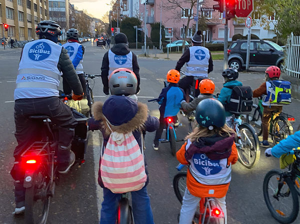 Foto: Gruppe von Kindern und Erwachsenen fährt gemeinsam mit Fahrrädern als Bicibus durch eine Stadtstraße.