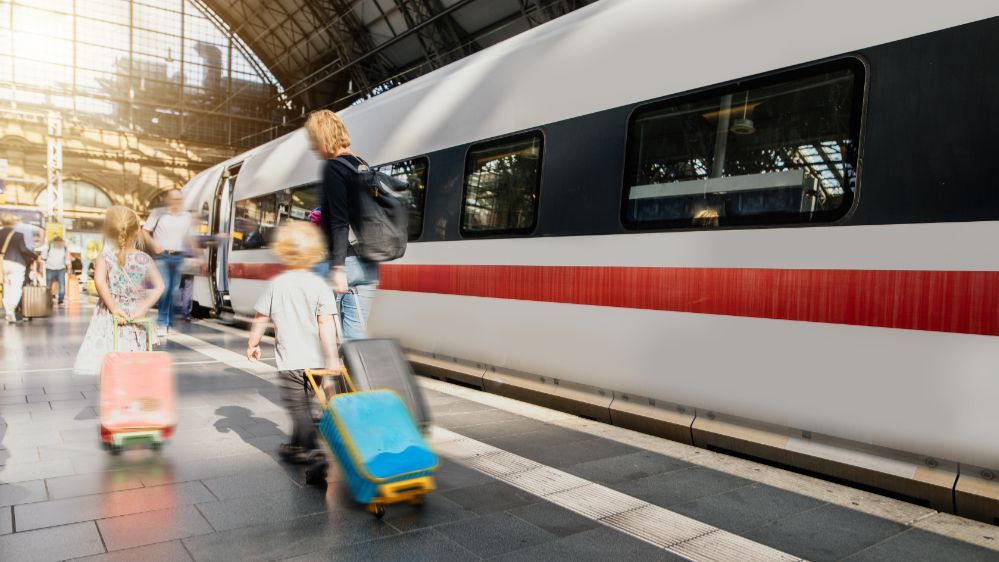 Mutter mit zwei kleinen Kindern steht am Bahngleis, während hinter ihnen ein ICE einfährt. Man sieht aus dem Bahnhof heraus das große Dach und die Sonne scheint herein