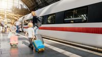 Mutter mit zwei kleinen Kindern steht am Bahngleis, während hinter ihnen ein ICE einfährt. Man sieht aus dem Bahnhof heraus das große Dach und die Sonne scheint herein