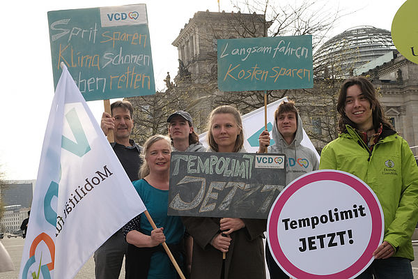 Das Bild zeigt eine Gruppe Hauptamtlicher des VCD bei einer gemeinsamen Aktion mit dem Tempolimit-Bündis. Die Personen stehen vorm Bundestag und halten VCD-Flaggen sowie Plakate mit der Aufschrift "Tempolimit jetzt!", "Langsam fahren, Kosten sparen" und "Sprit sparen, Klima schützen, Leben retten" hoch. Am rechten Bildrand steht eine Mitarbeiterin der Deutschen Umwelthilfe in grüner Jacke, die ebenfalls ein Schild mit der Aufschrift "Tempolimit JETZT!" in den Händen hält.