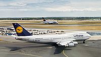 Das Bild zeigt eine Boeing 747 auf dem Rollfeld eines Flughafens. Im Hintergrund landet ein weiteres Flugzeug.