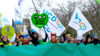 Das Bild zeigt einen Demozug von vorne, mit Schildern und Flaggen verschiedener Umweltverbände.