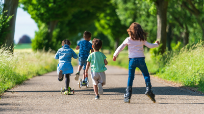Das Bild zeigt vier Kinder auf einem Feldweg von hinten. Eins der Kinder fährt auf Inlinern, zwei auf Tretrollern, eins rennt zu Fuß. In der Ferne sind unscharf Bäume und Felder zu erkennen.