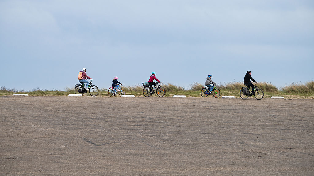 Foto: Gruppe von Radfahrenden fährt hintereinander über eine weitläufige, flache Landschaft unter bewölktem Himmel.
