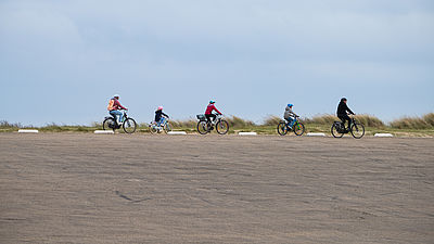 Foto: Gruppe von Radfahrenden fährt hintereinander über eine weitläufige, flache Landschaft unter bewölktem Himmel.