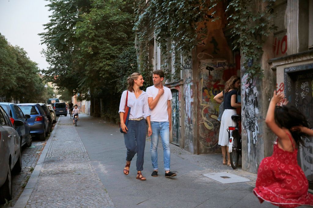 Eine Frau und ein Mann in sommerlichen Kleidern gehen einen Gehweg entlang und unterhalten sich. Der Gehweg ich sehr breit. Im Hintergrund fährt ein Kind Fahrrad, am rechten Rand tanzt ein Mädchen in einem roten Kleid. 