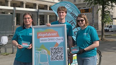 Das Bild zeigt drei Mitarbeitende des VCD in VCD-Shirts, wie sie mit Lastenrad und Beachflag ausgestattet an einer Berliner Bushaltestelle stehen. In den Händen halten sie Plakate bzw. Postkarten zur Bewerbung des VCD Mobilitätschecks ÖPNV.