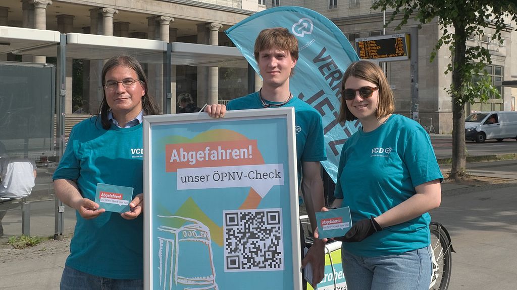 Das Bild zeigt drei Mitarbeitende des VCD in VCD-Shirts, wie sie mit Lastenrad und Beachflag ausgestattet an einer Berliner Bushaltestelle stehen. In den Händen halten sie Plakate bzw. Postkarten zur Bewerbung des VCD Mobilitätschecks ÖPNV.