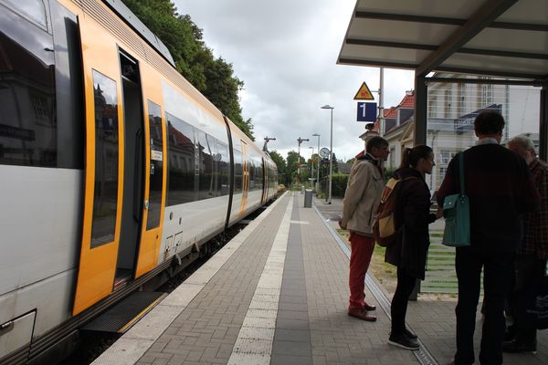 Man sieht den Bahnsteig am Regionalbahnhof Lübbecke (Westf.). Gerade steht eine Bahn dort, Menschen stehen in einem Grüppchen im Wartehäuschen.