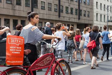 junge Frau schiebt Fahrrad im Schweigemarsch. Am Fahrrad klebt ein orangefarbenes Plakat auf dem steht: "3177 x für immer vermisst"