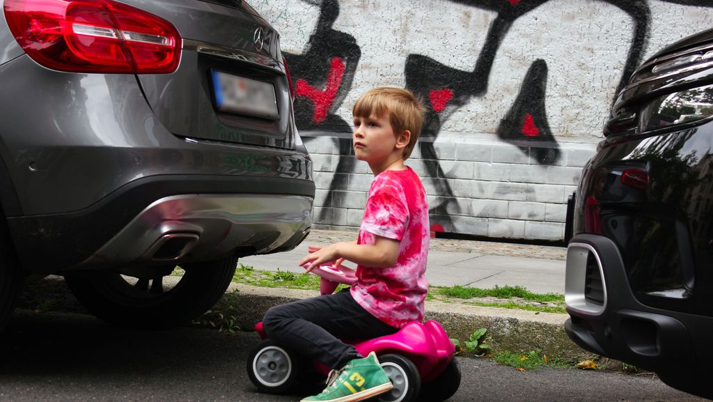 Kind auf rosa Bobbycar zwischen SUV / Autos