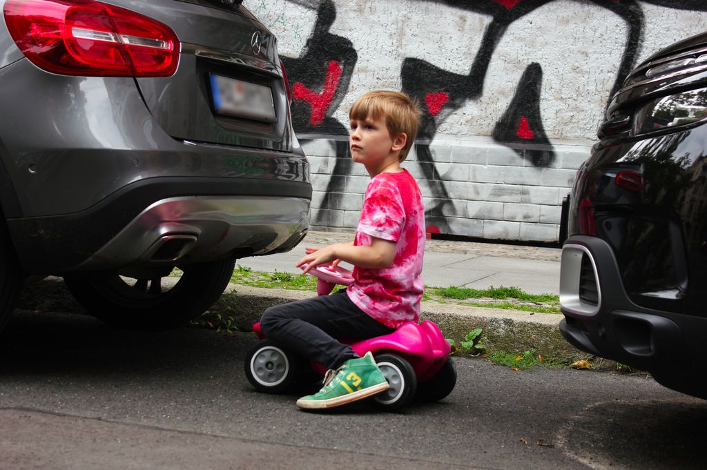 Kind auf rosa Bobbycar zwischen SUV / Autos