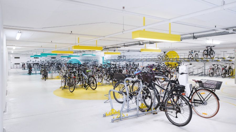 In einer sehr hellen, weiß gestrichenen Tiefgarage mit gelben Farbakzenten auf dem Boden, stehen viele Fahrräder in metallenen Ständen. Einige hängen auch an der Wand.