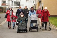 Eine Gruppe von Menschen, davon eine Frau mit Rollator, eine Frau mit Kinderwagen, eine Frau im Rollstuhl und eine Frau mit Fahrrad, bewegen sich eine Straße entlang.