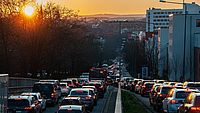Die Sonne geht über einer hügeligen Stadt unter. Im Vordergrund sieht man eine Straße mit Stau in beiden Richtungen.