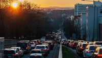 Die Sonne geht über einer hügeligen Stadt unter. Im Vordergrund sieht man eine Straße mit Stau in beiden Richtungen.