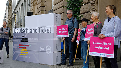 Das Bild zeigt eine VCD-Protestaktion zur Finanzierung des Deutschlandtickets aus dem Jahr 2025. Mehrere Personen stehen mit Plakaten neben einem Deutschland-Ticket-Aufsteller, der aus mehreren bedruckten und gestapelten Kartons besteht. Die Forderungen auf den Plakaten lauten: "Bus und Bahn ausbauen!" / "Bezahlbarer ÖPNV für alle!"