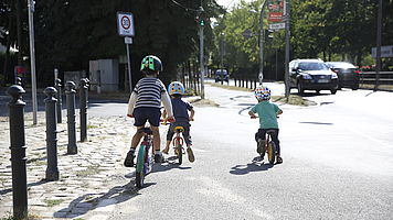Drei Kinder fahren mit ihren Fahrrädern bzw. Laufrädern am Rand einer vielbefahrenen Straße. Links von ihnen geht eine kleinere Straße ab, an der ein Tempo-30-Schild steht.