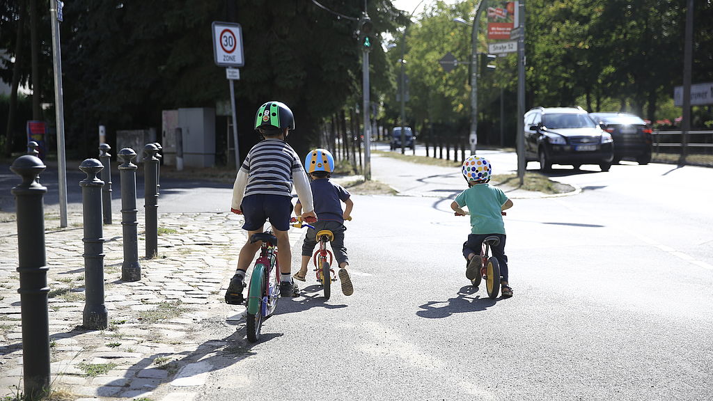 Drei Kinder fahren mit ihren Fahrrädern bzw. Laufrädern am Rand einer vielbefahrenen Straße. Links von ihnen geht eine kleinere Straße ab, an der ein Tempo-30-Schild steht.