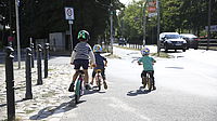 Drei Kinder fahren mit ihren Fahrrädern bzw. Laufrädern am Rand einer vielbefahrenen Straße. Links von ihnen geht eine kleinere Straße ab, an der ein Tempo-30-Schild steht.
