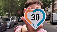 Eine Person steht auf einer Straße zwischen Bäumen und parkenden Autos und hält einen herzförmigen Tempo-30-Sticker des VCD in die Kamera.
