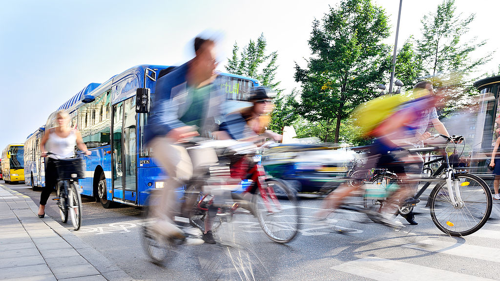 Foto: Radfahrende überqueren eine Straße an einer Haltestelle, im Hintergrund ein Bus.