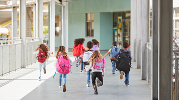 Das Bild zeigt mehrere Grundschulkinder mit Schulranzen, wie sie auf die Eingangstür eines Gebäudes zulaufen.