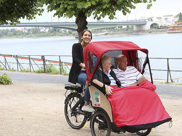 Foto: Eine Person fährt ein rotes Lastenrad mit überdachter Transportbox an einem Flussufer entlang.