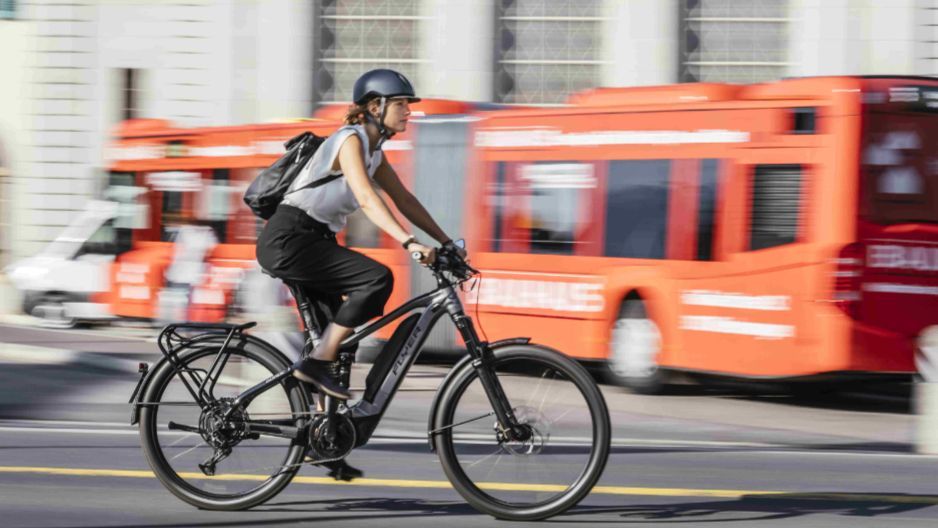 Foto zeigt eine Radfahrerin mit Helm, die auf einem S-Pedelec im Straßenverkehr unterwegs ist. Der Hintergrund ist leicht unscharf, dennoch erkennt man Autos und einen großen ÖPNV-Bus im Hintergrund des Geschehehens.
