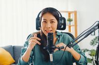 Eine junge asiatische Frau sitzt im Wohnzimmer am Schreibtisch vor einem Mikrofon.