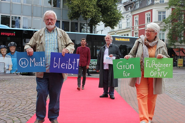 vier Menschen laufen auf einem roten Teppich auf die Kamera zu. Sie halten Schilder auf denen steht: "H Markt bleibt" und "Grün für Fußgänger & ÖPNV".