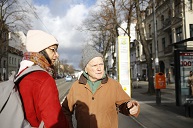 Ein älterer Mann erklärt einer jungen Frau etwas. Die beiden stehen auf der Straße an einer Straßenbahnhaltestelle. 