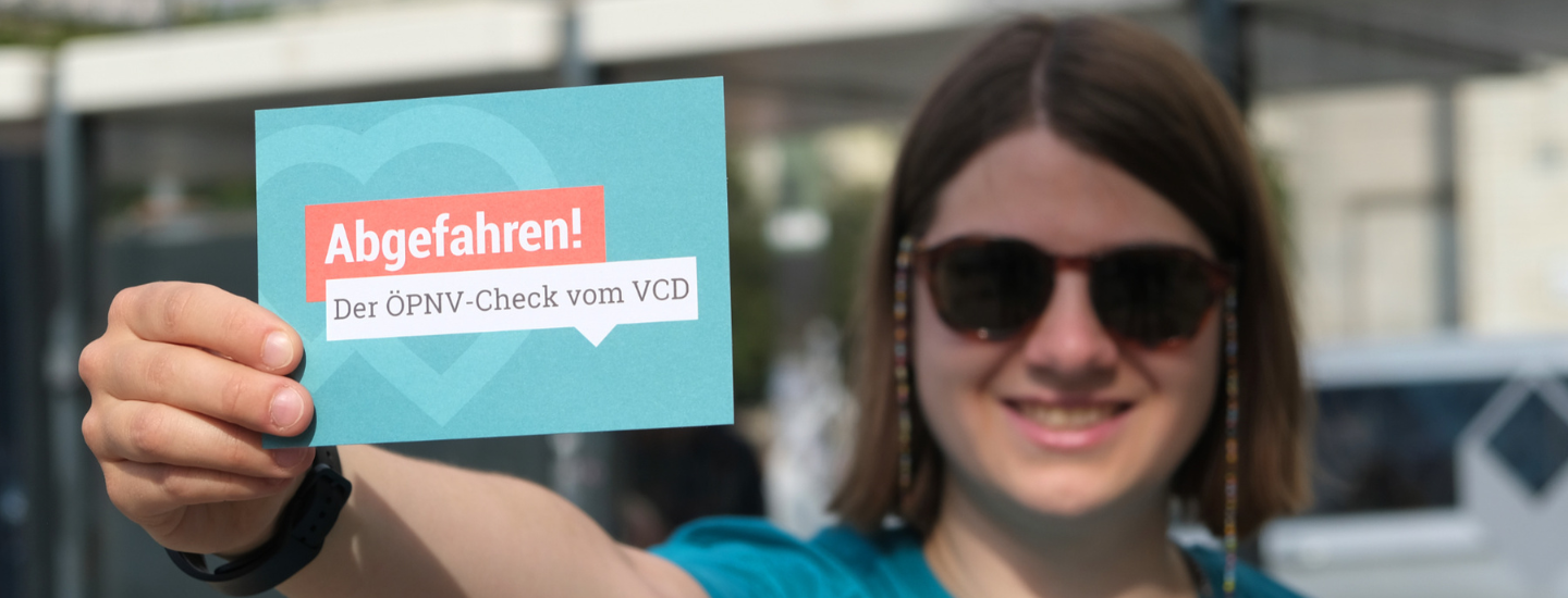Louise Beichler, Mitarbeiterin des VCD Bundesverbands, hält eine Postkarte zur Bewerbung des VCD-Mobilitätschecks ÖPNV in die Kamera. Sie trägt ein petrolfarbenes VCD-Shirt und eine Sonnenbrille. Im Hintergrund ist unscharf eine Bushaltestelle zu erkennen.