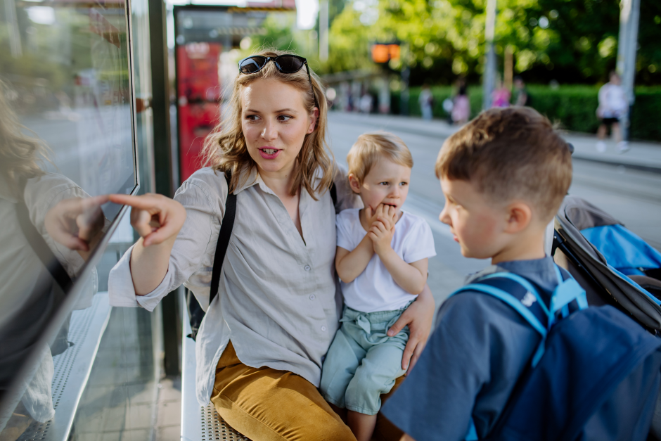 Weiblich gelesene Person mit zwei Kindern an einer Bushaltestelle