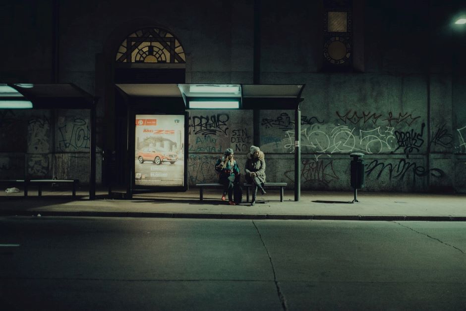 Zwei Menschen sitzen im Dunkeln an einer schwach ausgeleuchteten Bushaltestelle, im Hintergrund ist eine Wand mit Graffiti