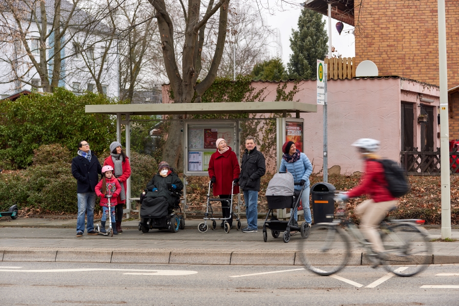 Menschen an Bushaltestelle: Rollstuhlfahrerin, Familie mit Kind, Frau mit Kinderwagen, älteres Pärchen mit Rollator, vorbei fährt Frau auf Fahrrad