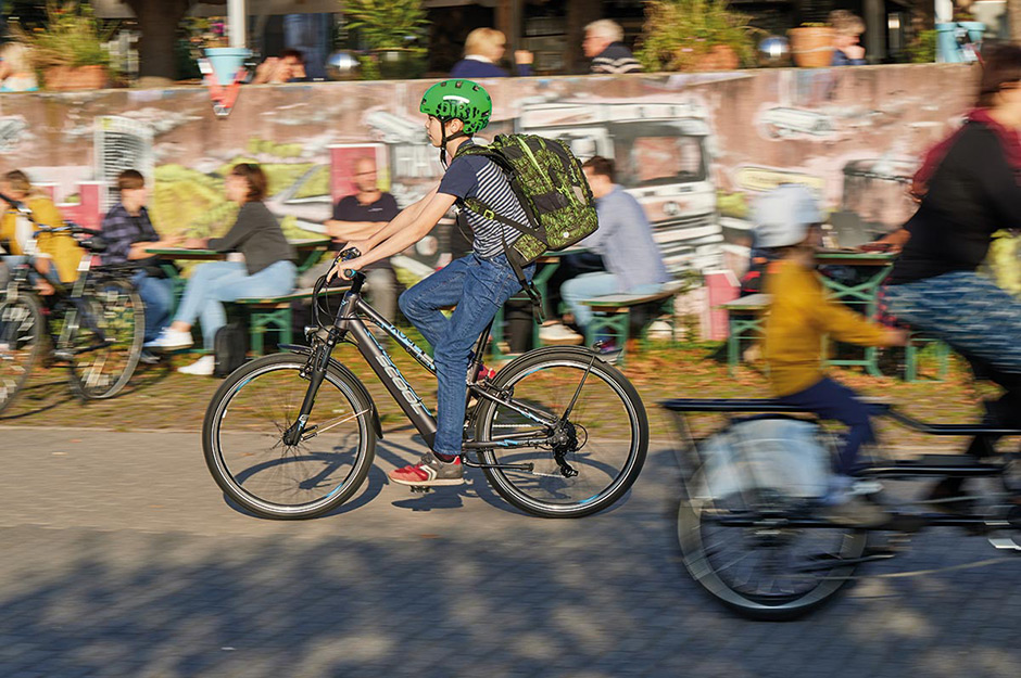 Ein Schuljunge mit grünem Fahrradhelm und Rucksack fährt auf seinem Fahrrad an einem Biergarten vorbei.