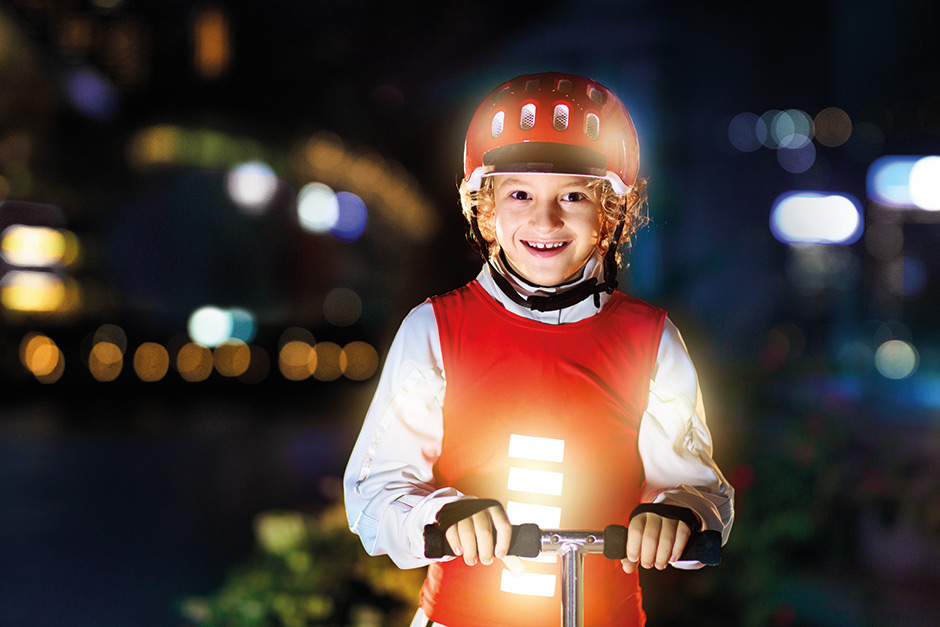 Kind mit einem beleuchteten roten Helm und einem Shirt mit Reflektoren fährt auf einem Roller durch die Dunkelheit.