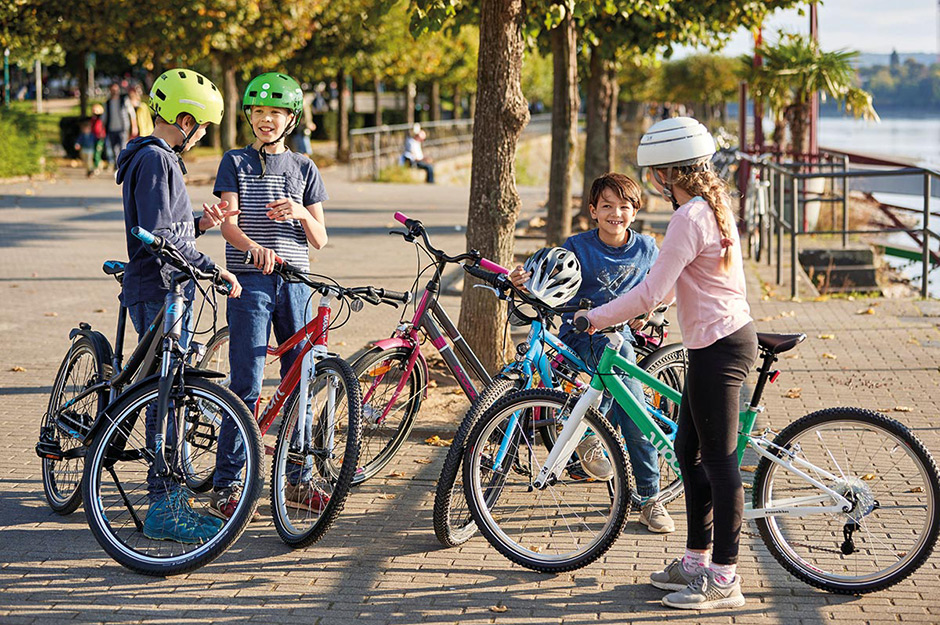 Vier Kinder, die Fahrradhelme tragen, stehen mit ihren Fahrrädern in Bonn an der Rheinpromenade und unterhalten sich.
